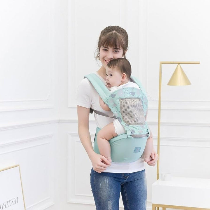 Woman holding a baby in a green baby carrier against a white wall.